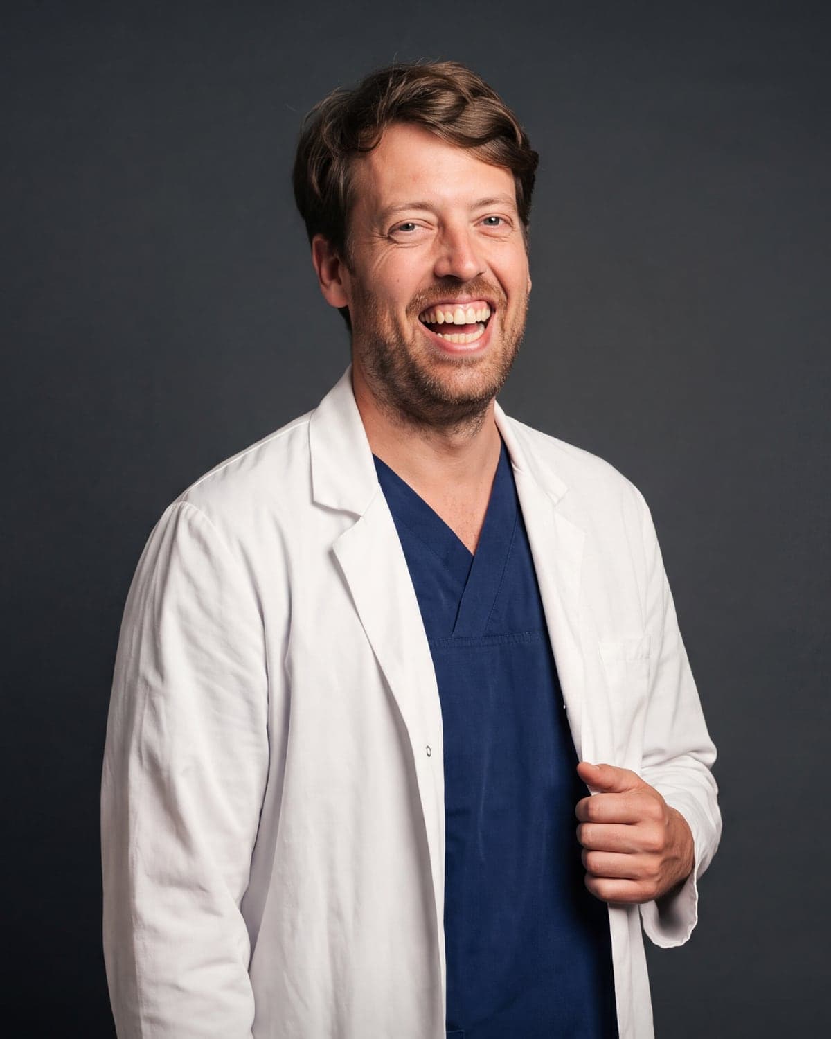 El Dr. Elio Díez-Feijóo, director médico de AdmiraVisión y adéu.now, riendo con bata blanca y pijama quirúrgico azul marino sobre un fondo oscuro de estudio