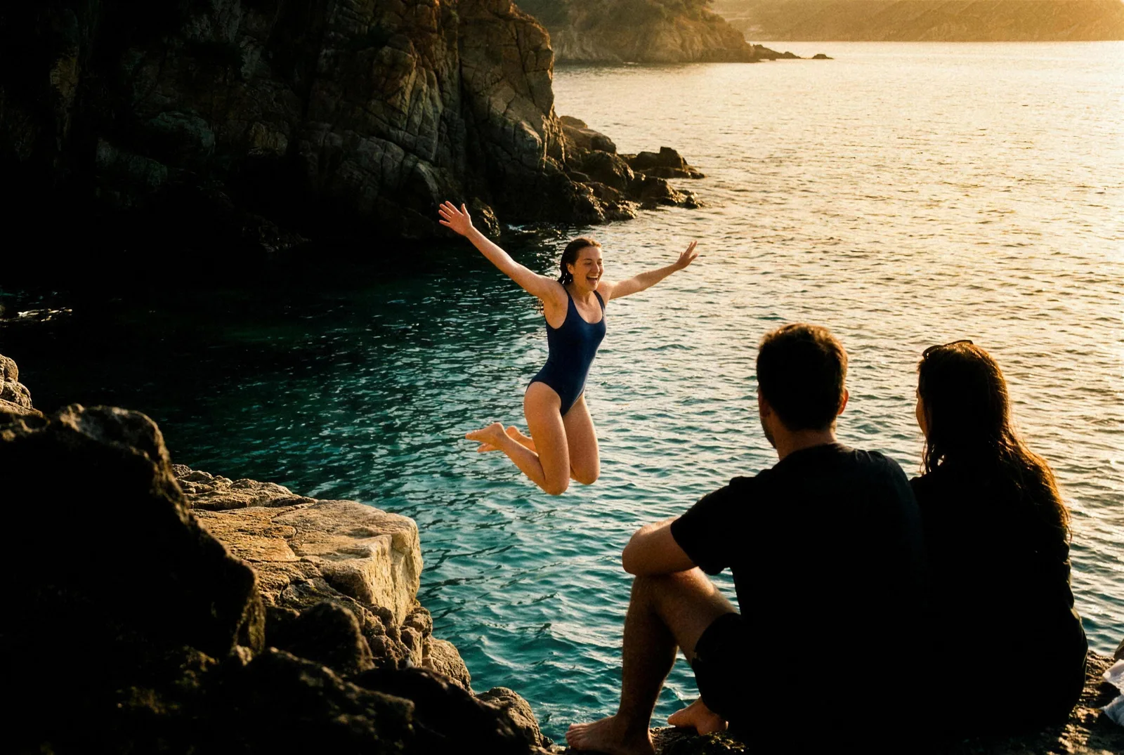 Una mujer salta al mar desde las rocas de una cala al atardecer mientras una pareja la mira sentada