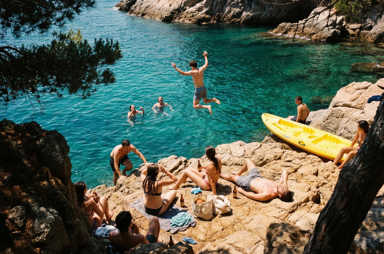 Amigos en una cala de rocas de la costa catalana: uno salta al mar turquesa, dos nadan y el resto toma el sol en las rocas